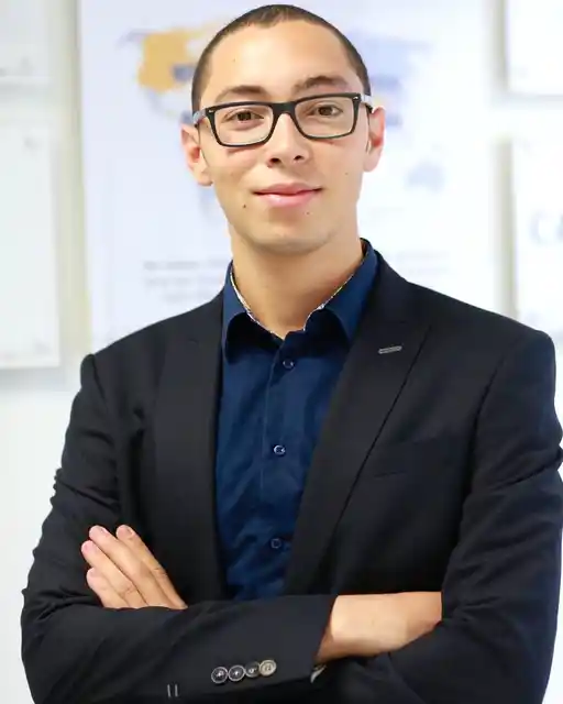 A person with glasses wearing a dark jacket and blue shirt, standing with folded arms.