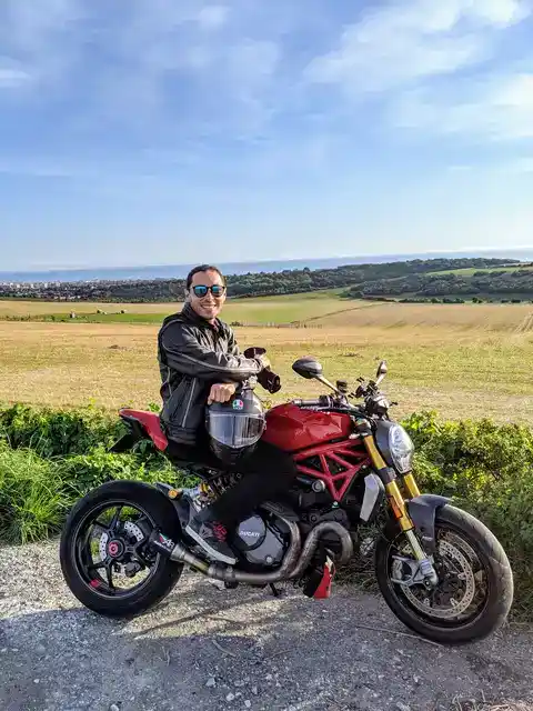 A rider on a red Ducati beside open fields and the sea.