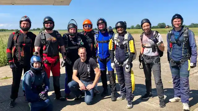 A group of skydivers wearing helmets and parachute harnesses at a sunny airfield.