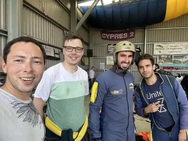 Four people gathered inside a skydiving hangar, with parachuting equipment and a canopy overhead.