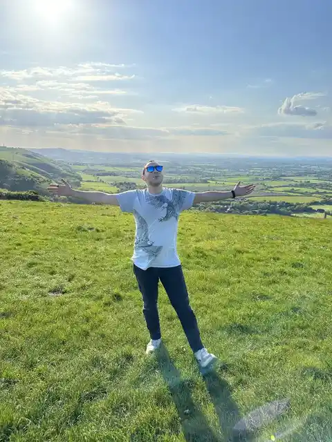 A person with outstretched arms on a grassy hillside overlooking fields under a bright sky.