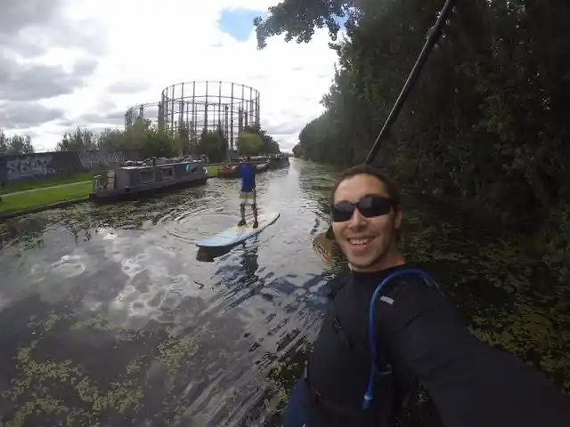 Two friends paddleboarding on a canal beside narrowboats and trees.