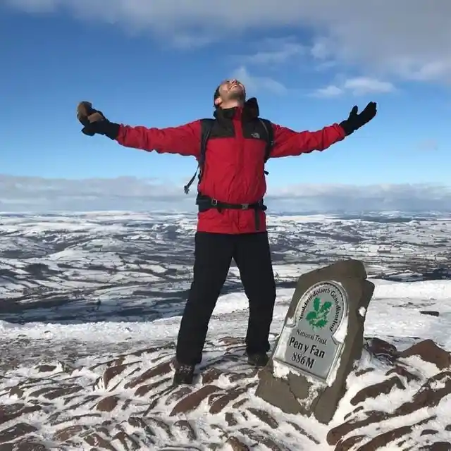 A hiker in a red jacket with arms outstretched beside the Pen y Fan summit marker in the snow.